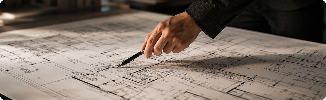 Hand hält Stift und zeigt auf detaillierten Architekturplan auf großem Tisch.